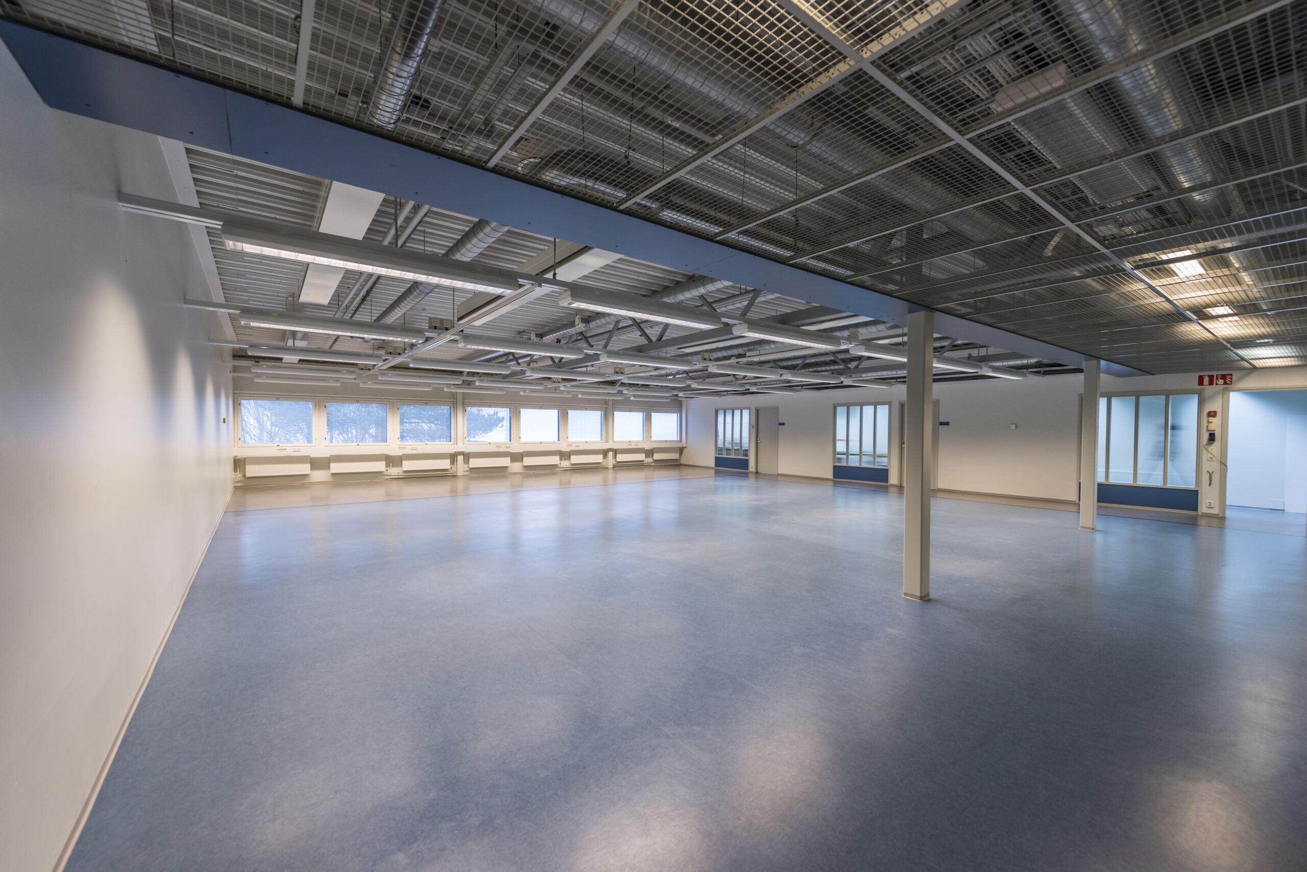Tyhjä tuotantotila, sininen muovipintainen lattia, katossa putkistoja ja teräsrakenteita. Paljon ikkunapinta-alaa.